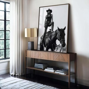 May include: A black and white framed photograph of a person in a cowboy hat and boots, standing on a horse. The photograph is displayed on a wooden console table with a gold lamp and books.