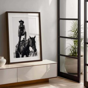 May include: Black and white framed print of a person in a cowboy hat and boots standing on a horse. The print is in a dark brown frame and is displayed on a white cabinet. A plant in a white pot is visible.