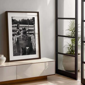 May include: Monochrome framed print featuring a woman in a cowboy hat, seated in a metal tub. The image includes cowboy boots, a fringed shawl, and a rural backdrop. The artwork is displayed on a white cabinet.