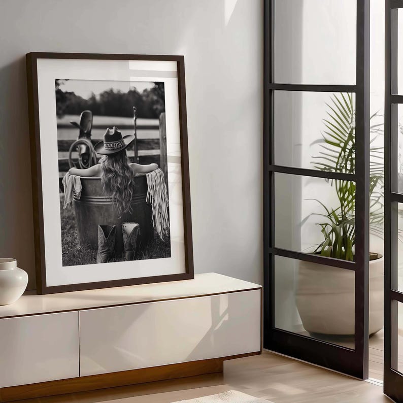 May include: Monochrome framed print featuring a woman in a cowboy hat, seated in a metal tub. The image includes cowboy boots, a fringed shawl, and a rural backdrop. The artwork is displayed on a white cabinet.