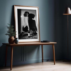 K&ouml;nnte beinhalten: Gerahmtes Schwarz-Wei&szlig;-Foto einer Frau mit Weinflasche und Glas. Das Foto steht auf einem Holztisch, im Hintergrund eine kleine Pflanze und eine Lampe. Der Rahmen ist braun.