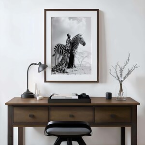 May include: Monochrome framed print featuring a person in a long dress posing with a zebra. The print is on a dark brown desk with a black lamp, candles, and a decorative tray.