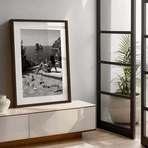 May include: A black and white framed wall art print depicting a person in a bikini sitting on a stone terrace overlooking the sea with rock formations and boats in the distance, with several pairs of high-heeled shoes scattered on the ground.