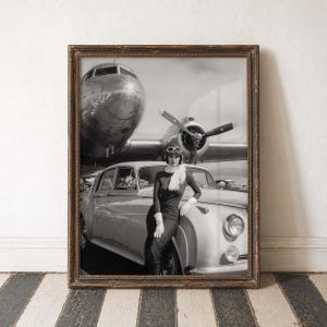 Op de afbeelding: Zwart-witfoto in een lijst met een vrouw in vintage kleding die poseert bij een klassieke auto, met een vliegtuig op de achtergrond. De foto zit in een bruine houten lijst, geplaatst tegen een gestreepte vloer en een witte muur.