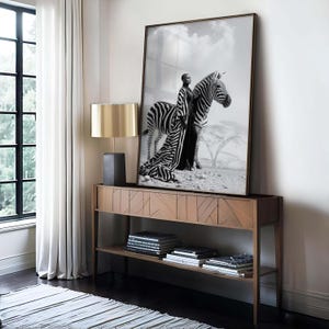 May include: A framed monochrome print of a person in a zebra-striped outfit standing beside a zebra. The print is displayed on a wooden console table, with a gold lamp.