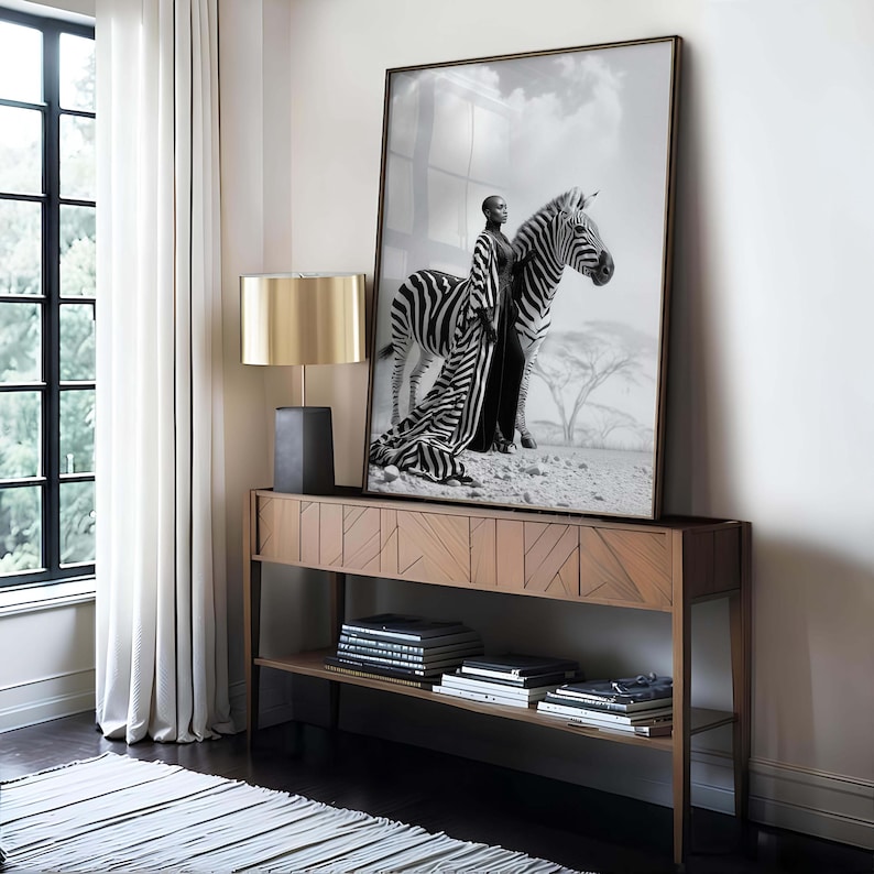 May include: A framed monochrome print of a person in a zebra-striped outfit standing beside a zebra. The print is displayed on a wooden console table, with a gold lamp.