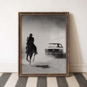 May include: Framed monochrome photograph depicting a cowboy on horseback and a classic Ford Mustang. The image shows the horse and car racing in a dusty environment. The frame is dark brown with gold detailing. The photograph is a vintage style.