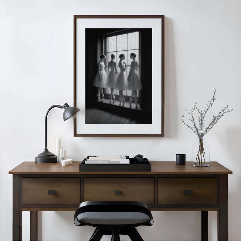 May include: A framed black and white photograph of four ballerinas in white tutus, standing by a window. The photograph is displayed above a wooden desk with a black lamp, candles, and a decorative tray.