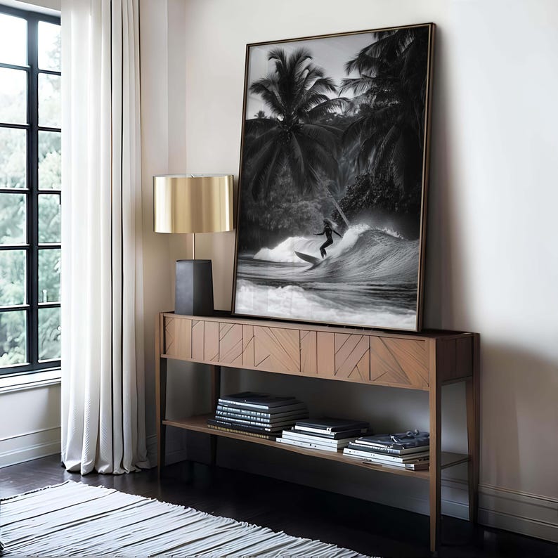 May include: A black and white framed print of a surfer riding a wave, set against a backdrop of palm trees. The artwork is displayed on a wooden console table with a gold lamp and books, creating a coastal theme.
