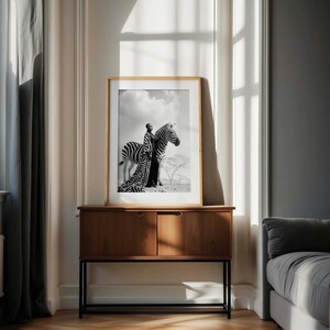 May include: Monochrome framed print depicting a person in a long dress with a zebra. The artwork is displayed on a wooden cabinet with a black metal frame. The room has neutral-toned walls and a window.