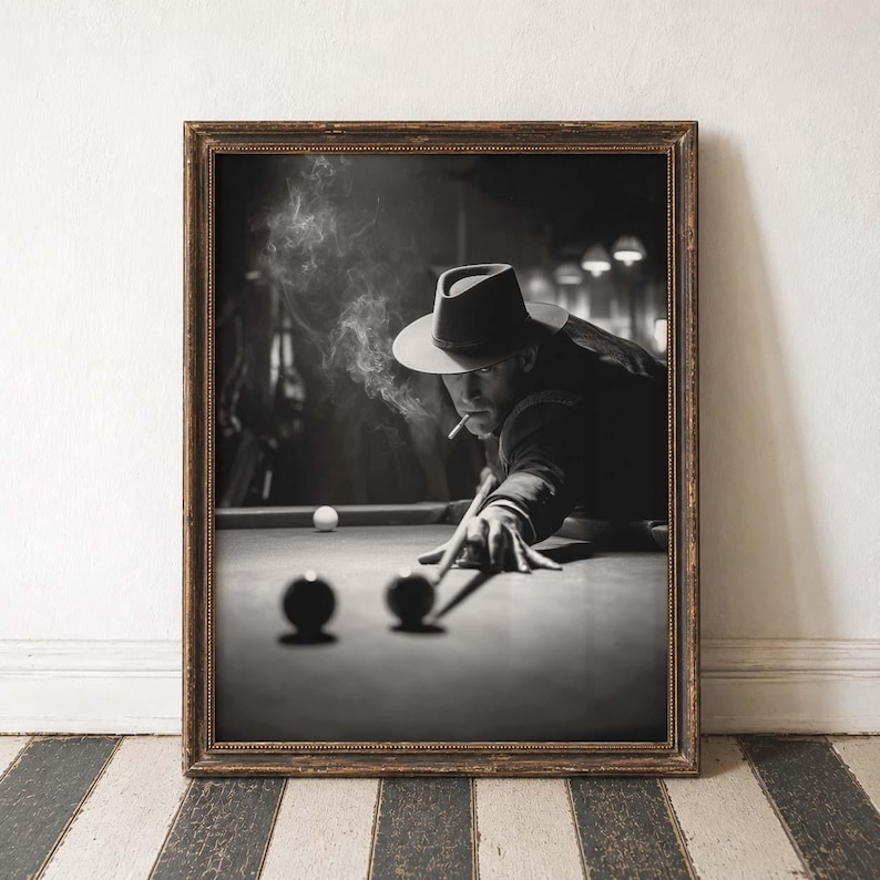 May include: Monochrome framed print depicting a person playing pool, smoking a cigarette. The person wears a fedora and is focused on the game. Smoke rises from the cigarette. Two pool balls are in the foreground.