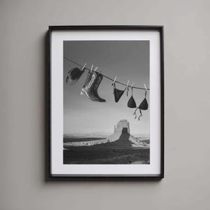 May include: Black and white framed print featuring a clothesline with a cowboy hat, cowboy boots, and a bikini hanging from it. The background shows a desert landscape with a large rock formation.