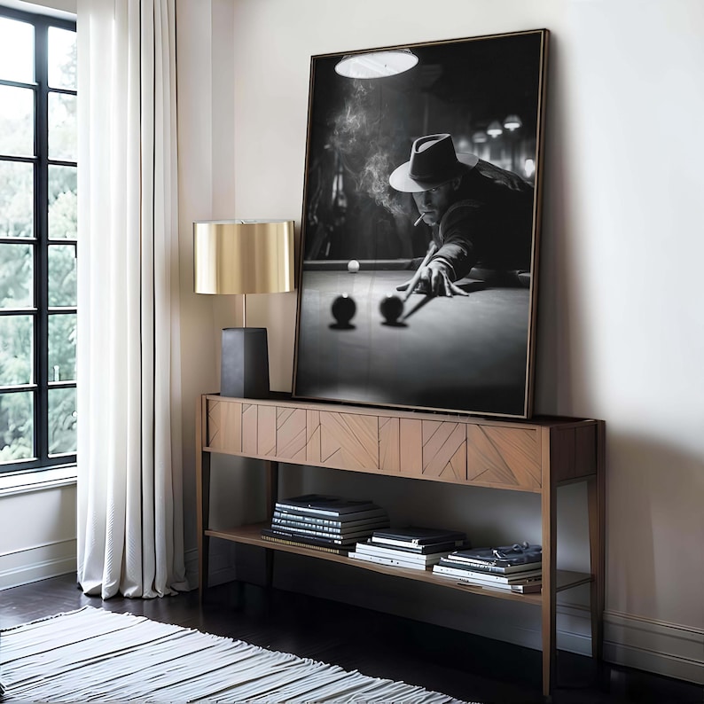 May include: Monochrome framed print of a person playing billiards, wearing a hat and smoking. The print is above a wooden console table with a gold lamp and books. The room has a window.