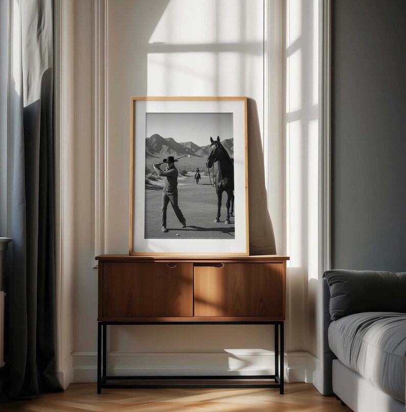May include: A framed black and white photograph of a golfer swinging a club, with a horse and mountains in the background. The photograph is displayed on a wooden cabinet with two doors and a black metal frame.