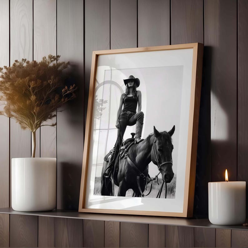 May include: Black and white framed print of a person in a cowboy hat and jeans standing on a horse. The print is in a light wood frame. A white candle and a potted plant are on the shelf.