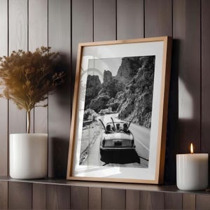 Puede incluir: Fotograf&iacute;a en blanco y negro enmarcada de un coche descapotable cl&aacute;sico en una carretera de monta&ntilde;a. El marco es de color marr&oacute;n claro. Una vela blanca y una planta en maceta est&aacute;n en el estante.