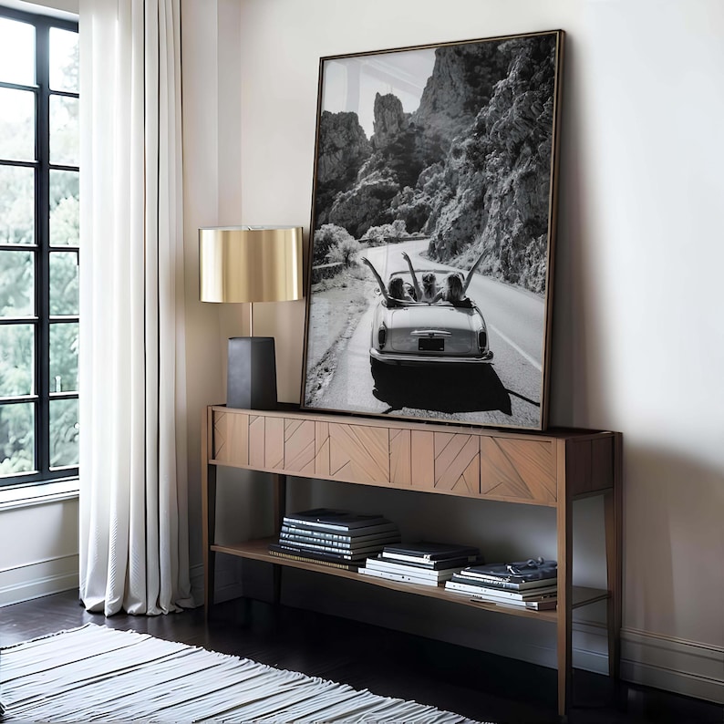 Puede incluir: Una fotograf&iacute;a en blanco y negro enmarcada de un coche descapotable vintage en una carretera sinuosa, expuesta sobre una consola de madera. Una l&aacute;mpara dorada con base negra est&aacute; a la izquierda. La habitaci&oacute;n tiene una ventana con cortinas blancas.
