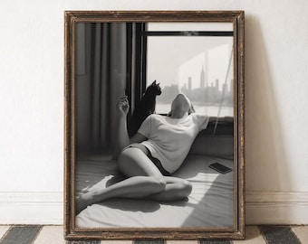 May include: Black and white framed print of a person reclining on a bed, smoking a cigarette, with a black cat and a city skyline visible through a window. The print is in a decorative brown frame.