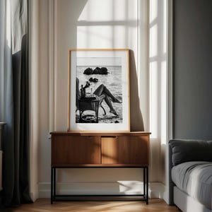 Pode incluir: Fotografia emoldurada a preto e branco de uma pessoa relaxando na praia, com uma mesa com uma garrafa e um copo. A fotografia &eacute; exibida em um arm&aacute;rio de madeira com duas portas e uma base de metal preta, contra uma parede neutra.