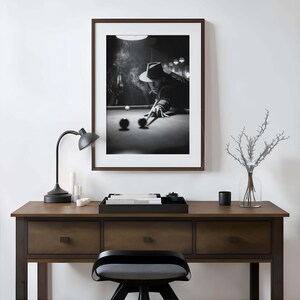 May include: Monochrome framed print depicting a person playing snooker, wearing a hat and smoking. The print hangs above a wooden desk with a black lamp, candles, and a tray. A black stool sits in front of the desk.