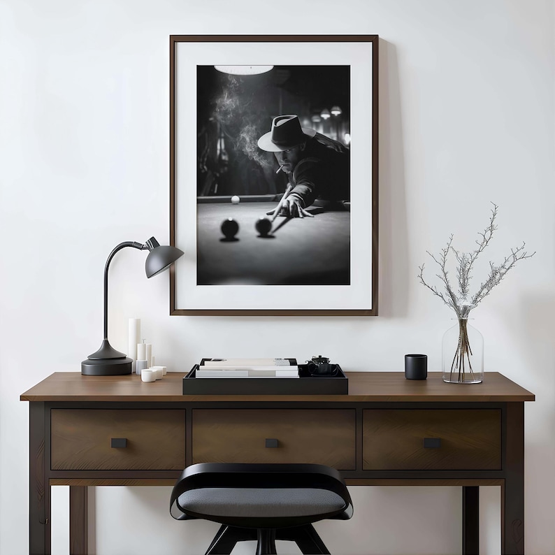 May include: Monochrome framed print depicting a person playing snooker, wearing a hat and smoking. The print hangs above a wooden desk with a black lamp, candles, and a tray. A black stool sits in front of the desk.