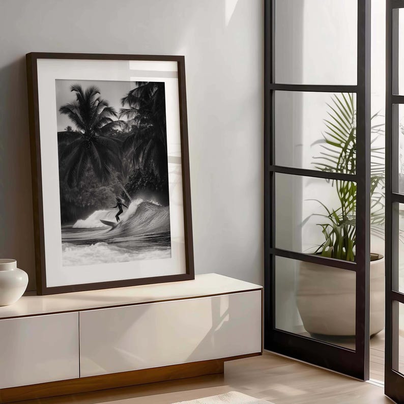 May include: Black and white framed print of a surfer riding a wave. The image features a surfer silhouetted against the wave, with palm trees in the background. The frame is dark brown, and the print is displayed on a white cabinet.