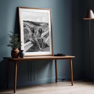 May include: A framed black and white photograph of two people sitting on a guardrail, one holding a sign that reads "TAKE ME ANYWHERE". The photograph is displayed on a wooden table with a plant and vase.