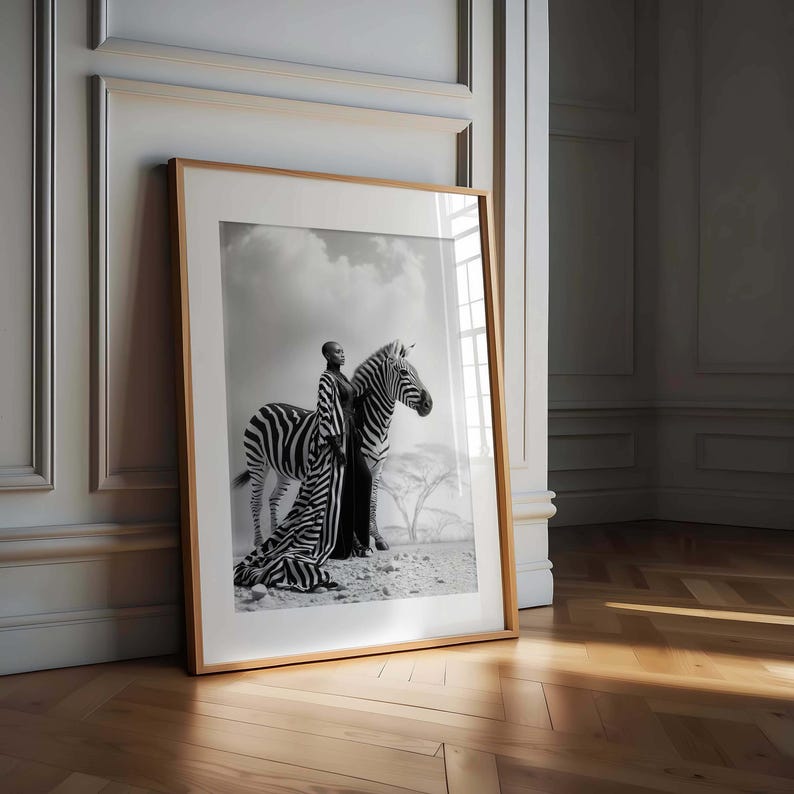 May include: Monochrome framed print depicting a person in a striped outfit alongside a zebra. The image is set against a backdrop of a cloudy sky and a desert scene. The frame is a light brown colour.