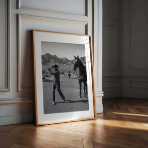 May include: Black and white framed print featuring a golfer swinging a club on a golf course. A horse stands nearby, and mountains are in the background. The print is in a wooden frame and leans against a wall.
