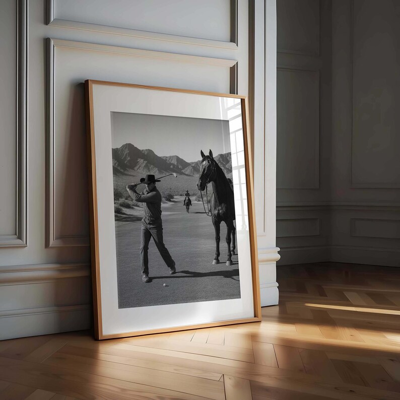 May include: Black and white framed print featuring a golfer swinging a club on a golf course. A horse stands nearby, and mountains are in the background. The print is in a wooden frame and leans against a wall.