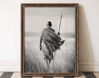 Op de afbeelding: Zwart-wit ingelijste print van een persoon die door hoog gras loopt en een speer vasthoudt. De persoon draagt een gestreept kledingstuk. De lijst is donker, versierd hout. De afbeelding staat tegen een neutrale achtergrond.