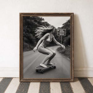 Peut inclure: Tirage encadr&eacute; en noir et blanc d'une personne faisant du skateboard sur une route. La personne porte un bikini et a de longs cheveux. Un panneau routier est visible en arri&egrave;re-plan. L'impression est dans un cadre marron.