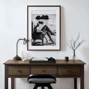 Pode incluir: Impress&atilde;o emoldurada em preto e branco de uma cena de praia. Um gramofone vintage est&aacute; em uma pequena mesa ao lado de uma cadeira de vime. A imagem apresenta uma garrafa, um copo e um disco de vinil.