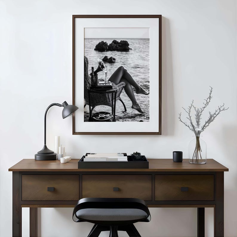 Pode incluir: Impress&atilde;o emoldurada em preto e branco de uma cena de praia. Um gramofone vintage est&aacute; em uma pequena mesa ao lado de uma cadeira de vime. A imagem apresenta uma garrafa, um copo e um disco de vinil.