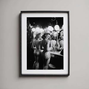 Może przedstawiać: Czarno-biała oprawiona grafika przedstawiająca dwie kobiety w błyszczących kostiumach, piórach i wyszukanych fryzurach, stojące przed lustrem. Obraz przedstawia scenę zza kulis w stylu vintage z jasnymi światłami i toaletką.