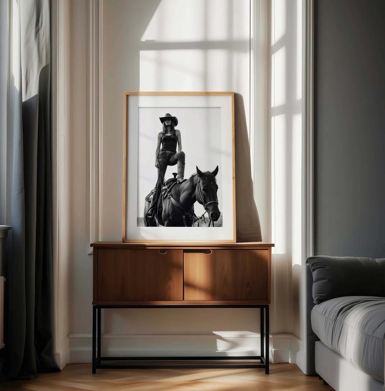 May include: Black and white framed print of a person in a cowboy hat and attire, standing on a horse. The artwork is displayed on a wooden cabinet with a black metal base, set against a neutral-toned wall and window.