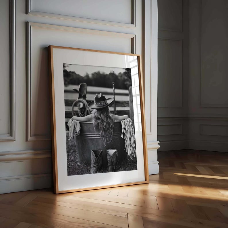 May include: Monochrome framed print depicting a person in a cowboy hat and boots, sitting in a metal tub. The image has a rustic, western theme, with a wooden fence and landscape in the background. The print is displayed in a wooden frame.