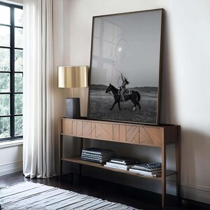 May include: A large framed black and white photograph of a person riding a horse and swinging a lasso, displayed on a wooden console table with a geometric front pattern, accompanied by a lamp with a metallic gold shade and stacked books.