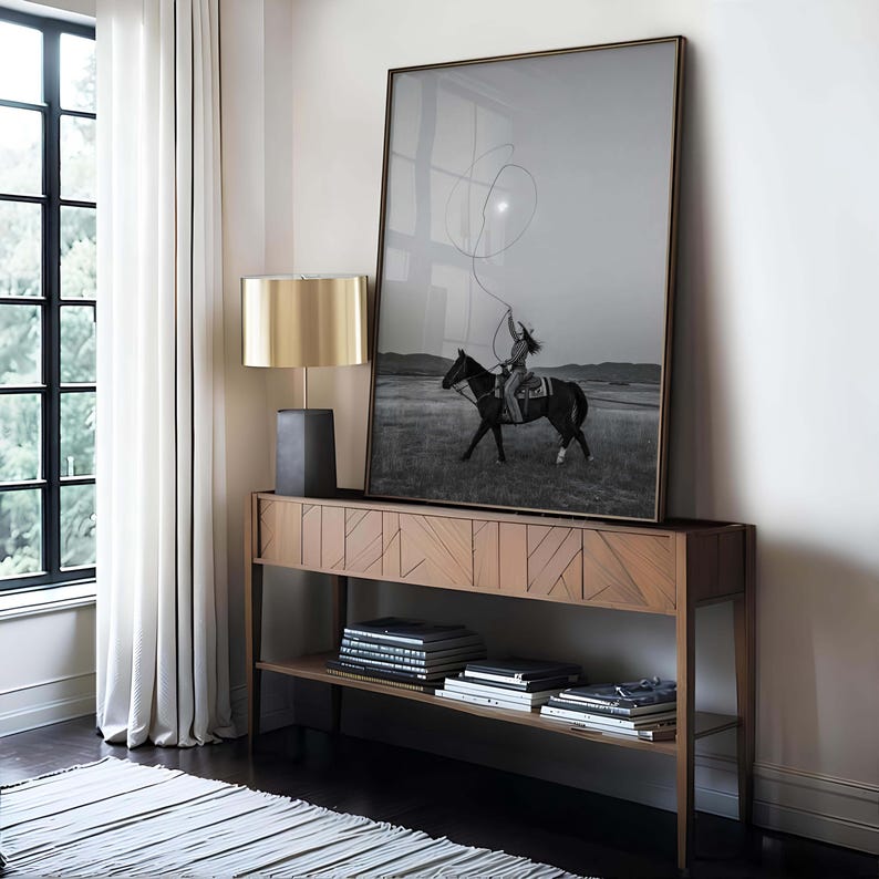 May include: A large framed black and white photograph of a person riding a horse and swinging a lasso, displayed on a wooden console table with a geometric front pattern, accompanied by a lamp with a metallic gold shade and stacked books.