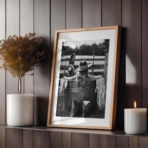 May include: Framed black and white photograph of a person in a cowboy hat and boots, relaxing in a metal tub. The image is set outdoors with a rustic fence in the background. A candle and a potted plant are on a shelf.