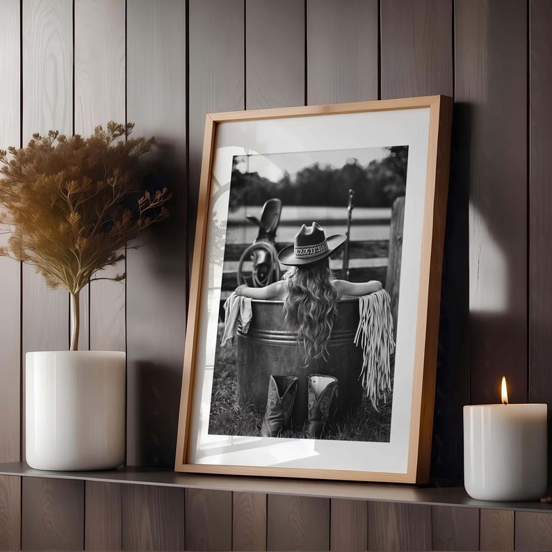 May include: Framed black and white photograph of a person in a cowboy hat and boots, relaxing in a metal tub. The image is set outdoors with a rustic fence in the background. A candle and a potted plant are on a shelf.