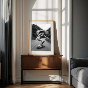 Peut inclure: Photographie en noir et blanc encadr&eacute;e d'une personne faisant du skateboard sur une route. L'image est pr&eacute;sent&eacute;e sur une commode en bois avec un cadre en m&eacute;tal noir. La pi&egrave;ce a une fen&ecirc;tre avec de la lumi&egrave;re naturelle et un rideau gris.