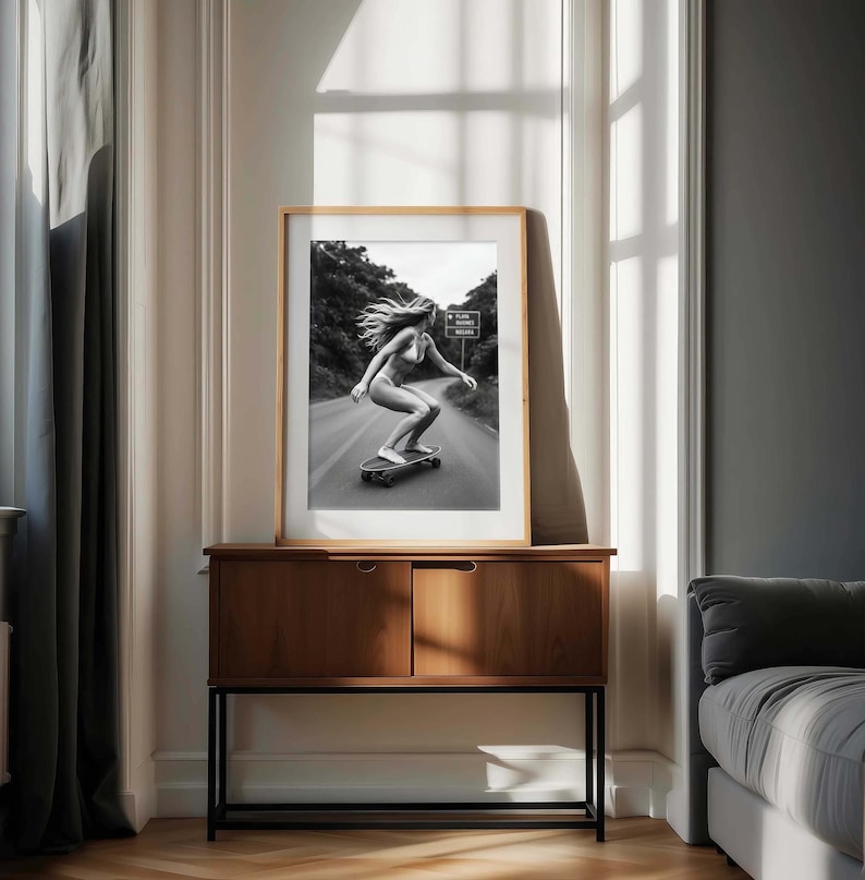 Peut inclure: Photographie en noir et blanc encadr&eacute;e d'une personne faisant du skateboard sur une route. L'image est pr&eacute;sent&eacute;e sur une commode en bois avec un cadre en m&eacute;tal noir. La pi&egrave;ce a une fen&ecirc;tre avec de la lumi&egrave;re naturelle et un rideau gris.