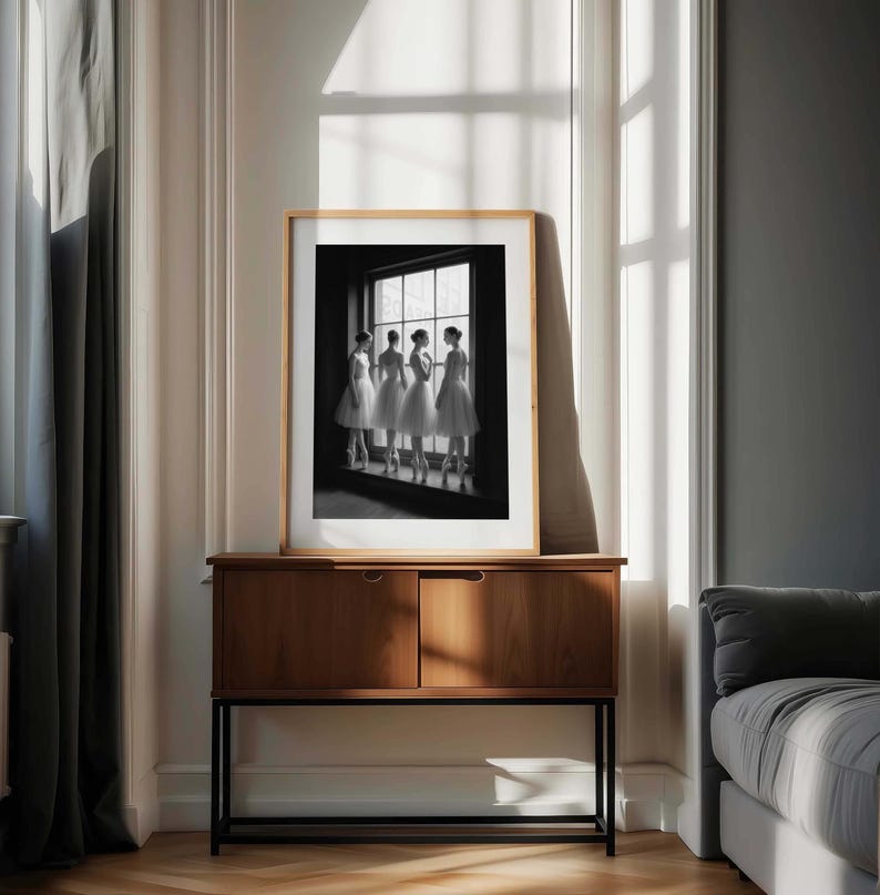 May include: A framed black and white photograph of five ballerinas in white tutus, standing by a window. The photograph is displayed on a wooden cabinet with a black metal frame, set against a neutral-toned wall with a window and curtains.