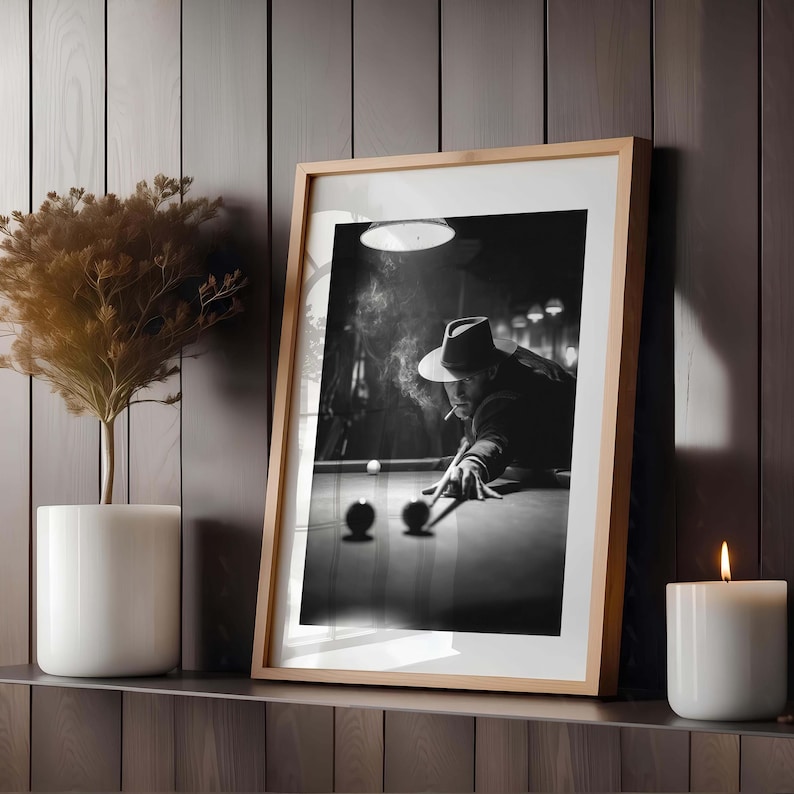 May include: Monochrome framed print of a man playing billiards, wearing a hat and smoking. The image is in a wooden frame, and the scene is lit by a single overhead light. A candle and a plant are on the shelf.