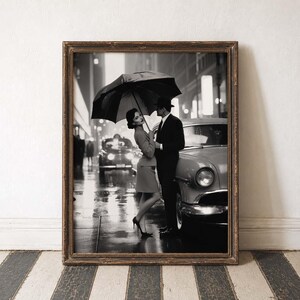 May include: Black and white framed print of a couple under an umbrella in the rain next to a classic car. The woman wears a skirt suit and heels, while the man is in a suit and hat. The background shows city lights and other cars.