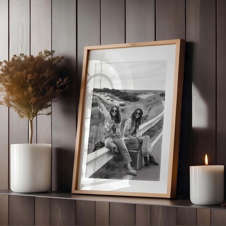 May include: Black and white framed photograph of two people sitting on a guardrail, holding a sign that says "TAKE ME ANYWHERE". The photograph is in a light wood frame, and is displayed on a shelf with a white candle and a decorative plant.