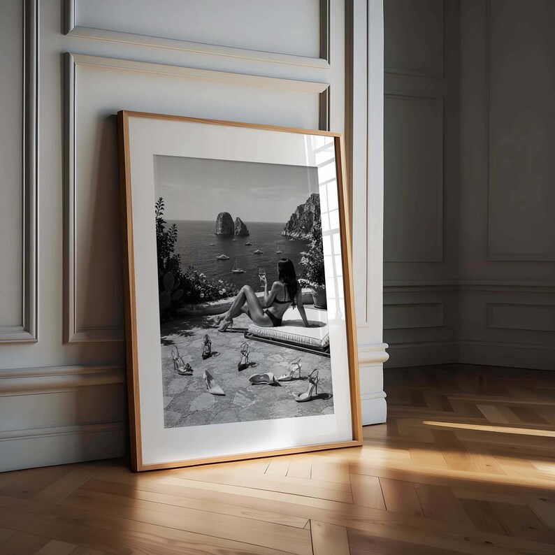 May include: A framed black and white photograph depicting a person in a bikini sitting on a lounge chair on a stone terrace overlooking the ocean with rocky cliffs in the distance. Several pairs of high-heeled shoes are scattered on the ground.