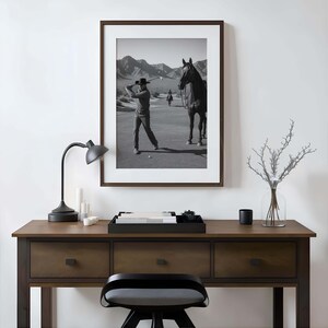 May include: Black and white framed print of a golfer swinging a club on a golf course, with a horse standing nearby. The print is displayed above a wooden desk with a black lamp, candles, and a tray.