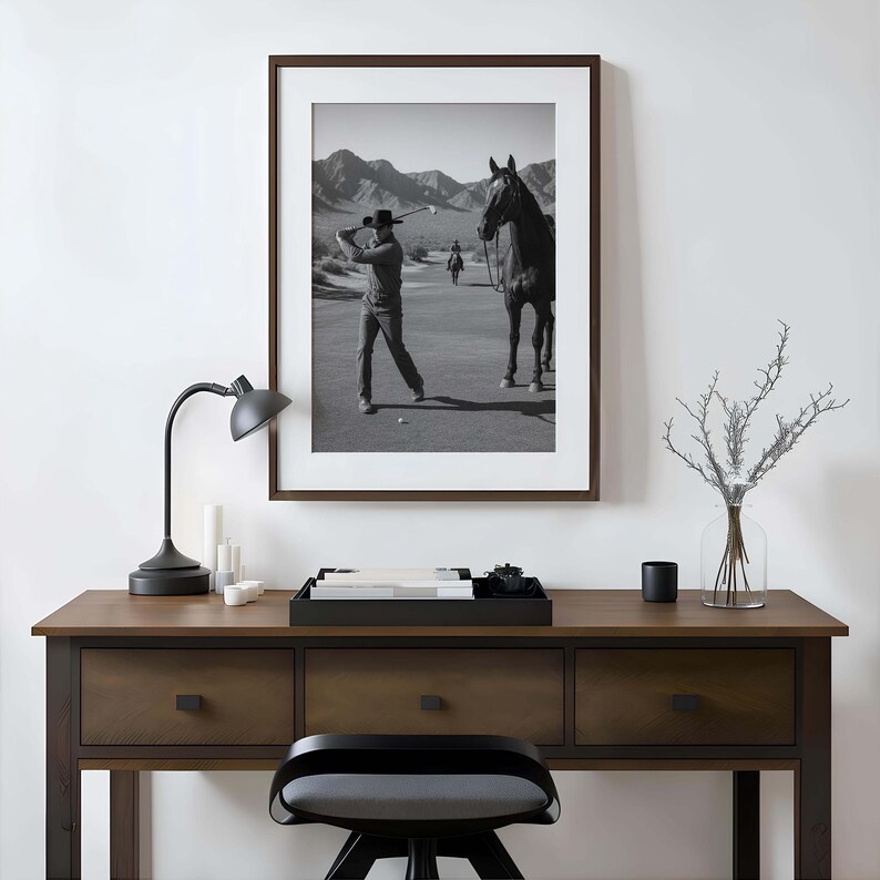 May include: Black and white framed print of a golfer swinging a club on a golf course, with a horse standing nearby. The print is displayed above a wooden desk with a black lamp, candles, and a tray.